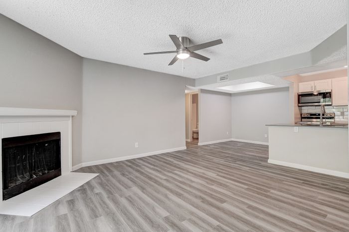 Spacious living room featuring elegant wood flooring, a modern ceiling fan, and a cozy fireplace, flowing into an open kitchen with sleek appliances.