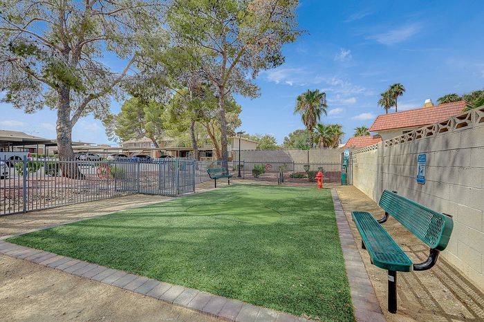 Charming outdoor space featuring a fenced dog park with lush green grass, benches, and scenic trees under a blue sky.