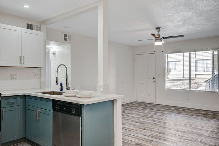 Modern open-concept kitchen with teal cabinets and stainless steel appliances, leading to a bright living area with elegant wooden flooring and large windows.