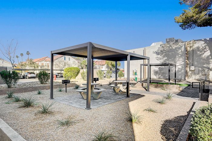 Charming outdoor space with modern pergola, picnic tables, and a built-in grill set against a backdrop of lush greenery and clear blue skies.