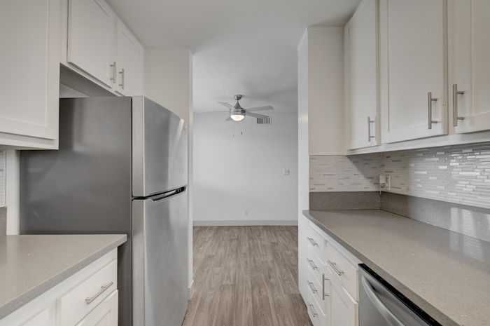 Sleek modern kitchen with stainless steel fridge, elegant cabinetry, and stylish backsplash, opening into a bright living space with a ceiling fan.