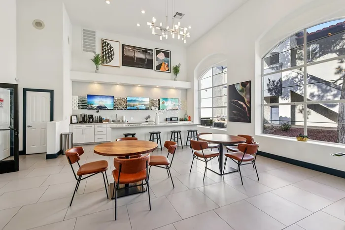 Modern community lounge with high ceilings, stylish chandeliers, and large windows, featuring cozy seating and a sleek kitchenette with vibrant artwork.