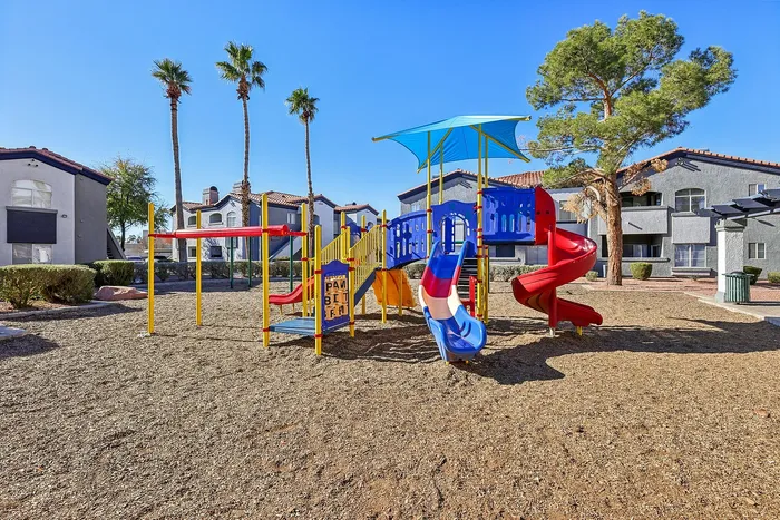 Vibrant playground with colorful slides and climbing structures, surrounded by lush palm trees and modern apartment buildings.