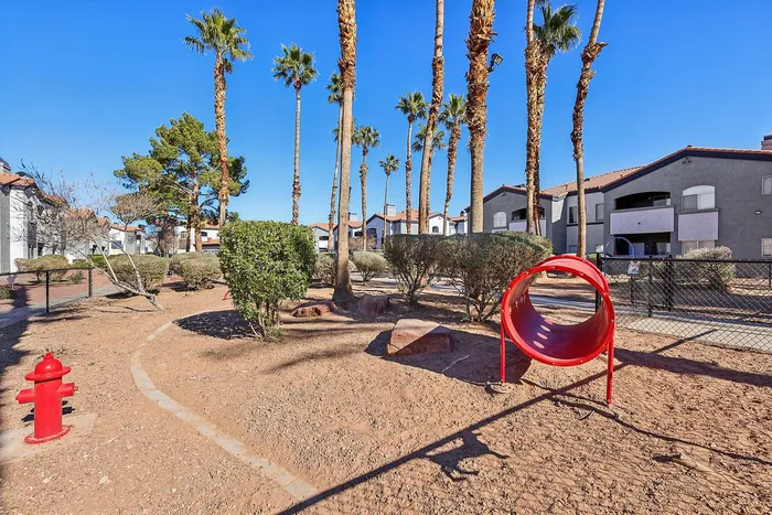 Inviting outdoor play area with red play tunnel, surrounded by palm trees and modern apartment buildings under clear blue skies.