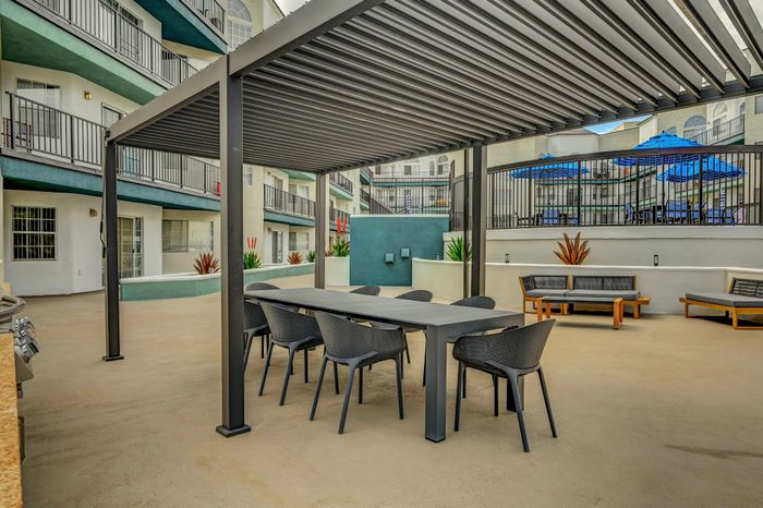 Modern outdoor courtyard with stylish seating area, sleek dining table, and vibrant blue umbrella shades, surrounded by elegant apartment balconies.