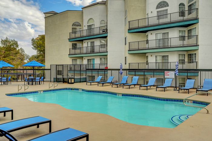 Inviting apartment complex featuring a sparkling swimming pool surrounded by modern lounge chairs, with a backdrop of stylish balconies and lush trees.