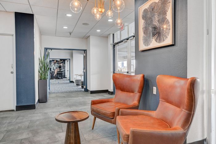 Inviting lobby with stylish leather chairs, elegant chandelier lighting, and modern artwork, offering a glimpse into a chic fitness area in the background.