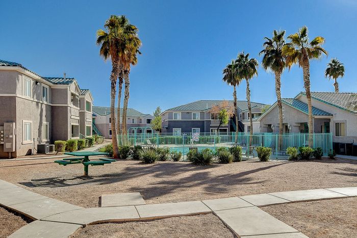 Beautiful apartment complex featuring modern buildings, lush palm trees, and a refreshing outdoor pool surrounded by green fencing.