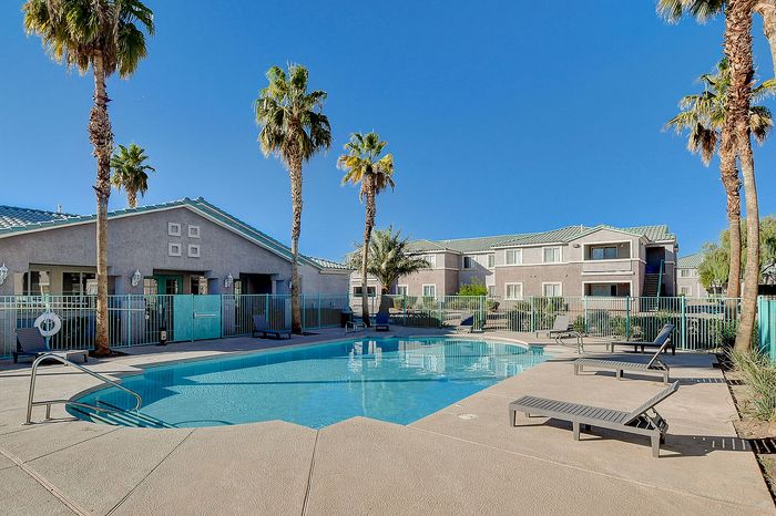 Vibrant apartment complex featuring a sparkling pool surrounded by tropical palm trees and modern lounging chairs under a clear blue sky.