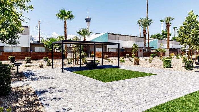 Modern apartment complex with beautifully landscaped grounds featuring palm trees, a shaded seating area, and a barbecue space under a clear blue sky.