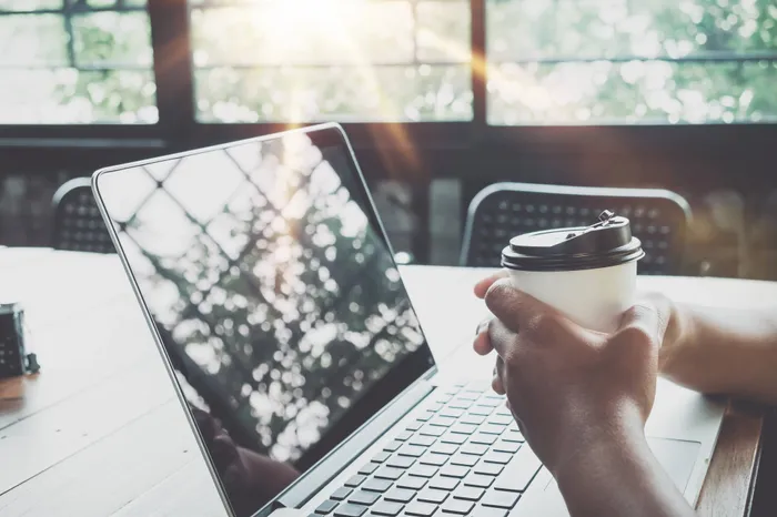 Stock photograph of someone drinking a coffee and sitting at their computer. Light is shining through the window.
