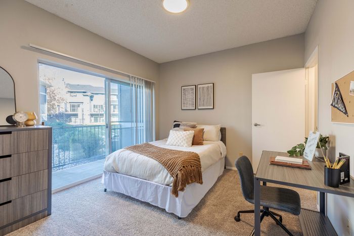 Model bedroom with white walls, tan carpet, large glass sliding door leading to a balcony and furniture pieces including a full size bed, dresser, desk, and chair