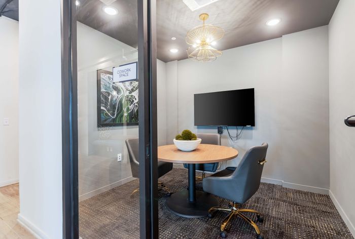 Modern coworking space with sleek decor, featuring a round wooden table, comfortable chairs, stylish lighting, and a mounted TV.