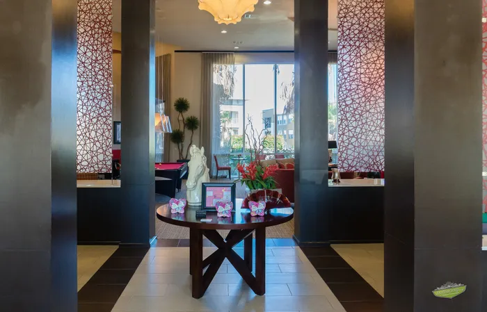 Elegant and modern lobby with stylish decor features, a round wooden table adorned with decorative butterflies and vibrant plants. Sunlight streams in through large windows, illuminating the open, welcoming space.