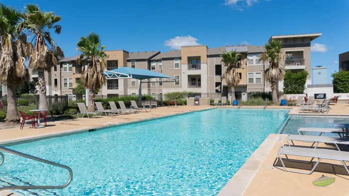 Luxurious apartment complex with a pristine swimming pool, surrounded by palm trees and ample lounge seating, under a clear blue sky.