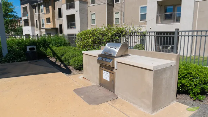 Modern outdoor BBQ area with a sleek stainless steel grill set against lush greenery and contemporary apartment buildings.
