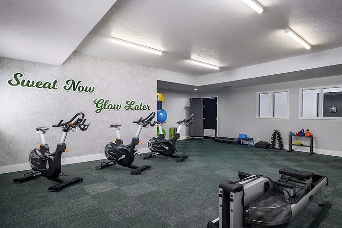 Fitness room with bikes and motivational wall art 'Sweat Now, Glow Later' at Campus Village apartments close to MSU.