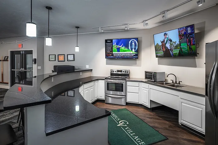 Modern communal kitchen with sleek countertops, stainless steel appliances, dual flat-screen TVs, and stylish pendant lighting.