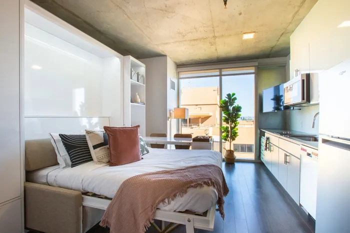Modern studio apartment with cozy bed, stylish kitchenette, and abundant natural light from large windows.