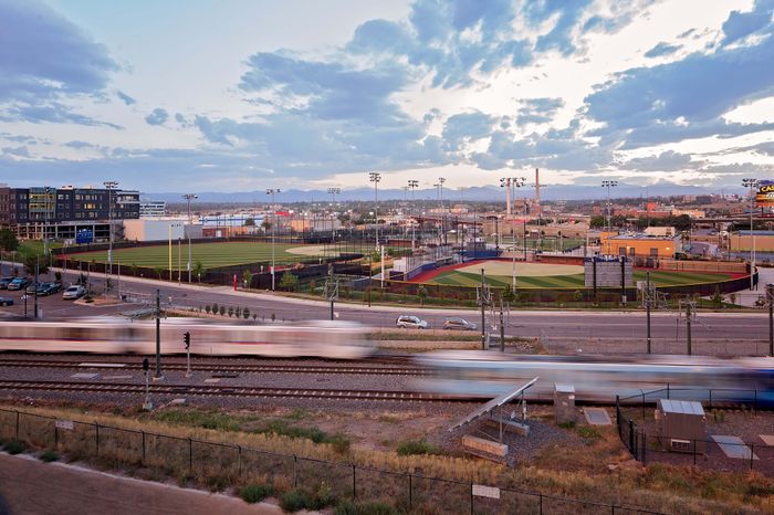 Vibrant urban landscape with a baseball field, modern buildings, and a sleek train speeding by, set against a stunning mountain backdrop.