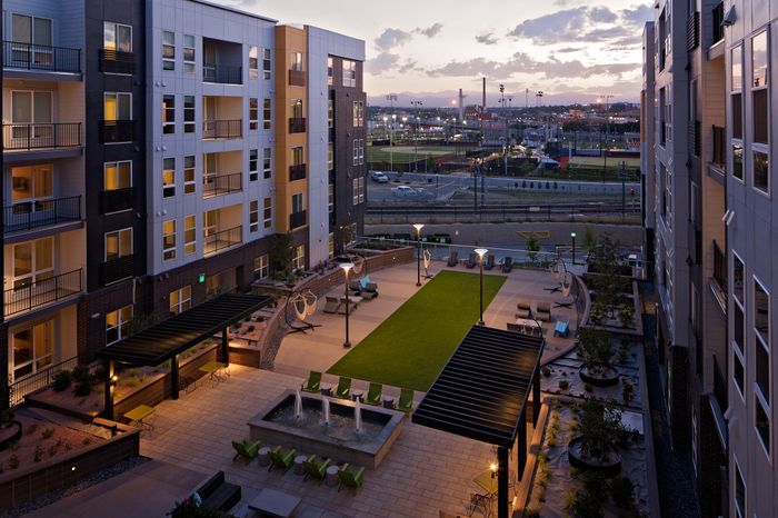 Evening view of a modern apartment complex with vibrant outdoor courtyard, featuring lounge chairs, greenery, and decorative lighting.