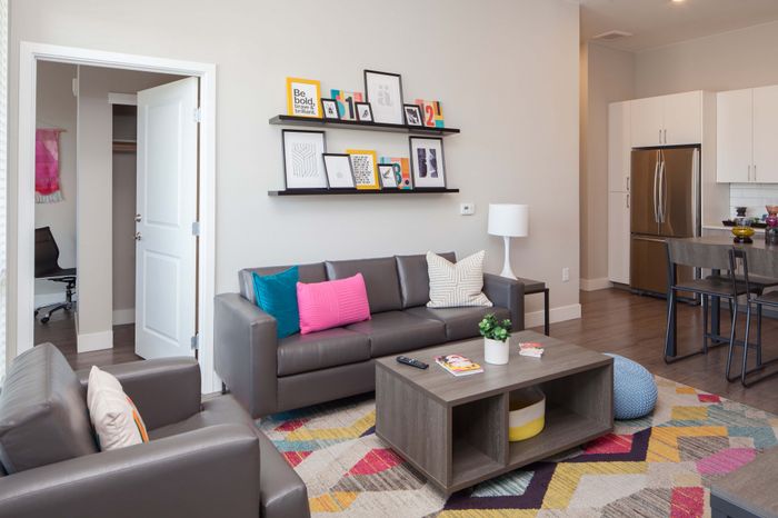 Modern living room with sleek gray sofas, vibrant cushions, and chic wall art, adjacent to a stylish kitchen with stainless steel appliances.