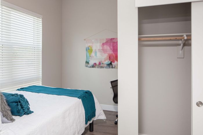 Bright and cozy bedroom featuring a comfortable white bed accented with teal throws, ample closet space, and a modern work chair beneath vibrant wall art.