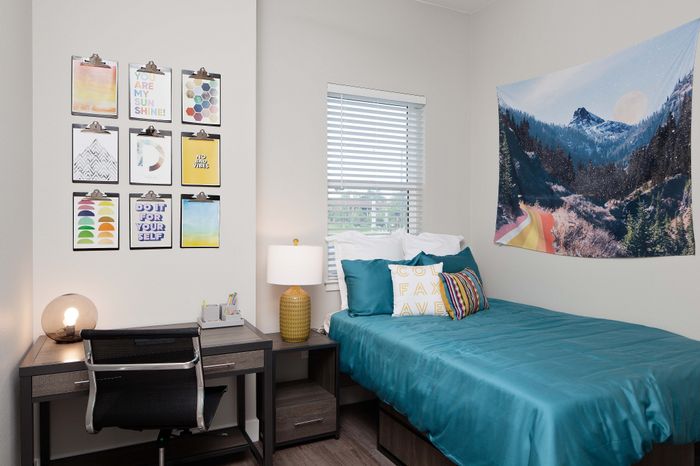 Cozy bedroom with vibrant decor, featuring a teal bedspread, artistic wall hangings, and a stylish workspace, perfect for modern living.