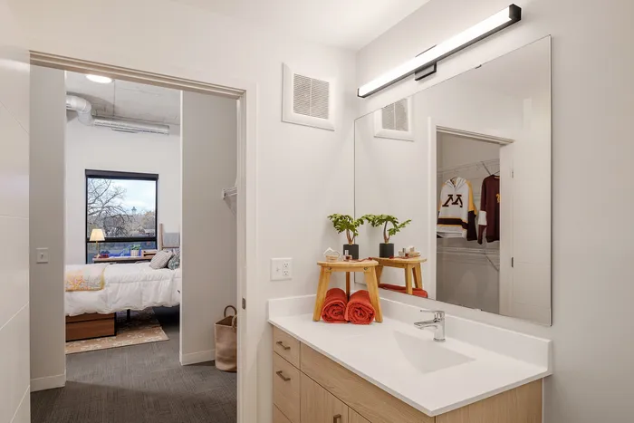 Chic apartment with a modern bathroom and cozy bedroom view, featuring a well-lit vanity, elegant mirror, and inviting bedroom decor.