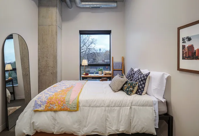 Cozy bedroom with a plush bed, vibrant pillows, and a large window offering a serene view, complemented by a stylish floor-length mirror and a chic desk lamp.