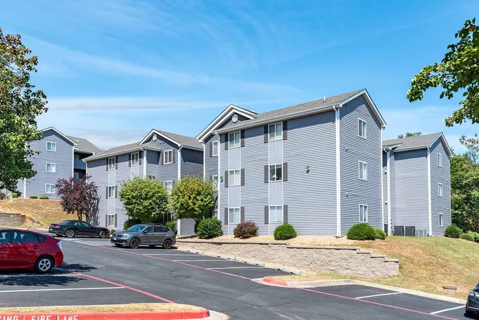 Charming modern apartment complex with stylish blue siding and well-groomed landscaping, offering convenient parking and lush greenery under a bright blue sky.