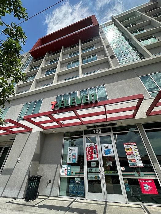Modern urban building with large glass windows, featuring a 7-Eleven store entrance and a vibrant red architectural accent, under a clear blue sky.