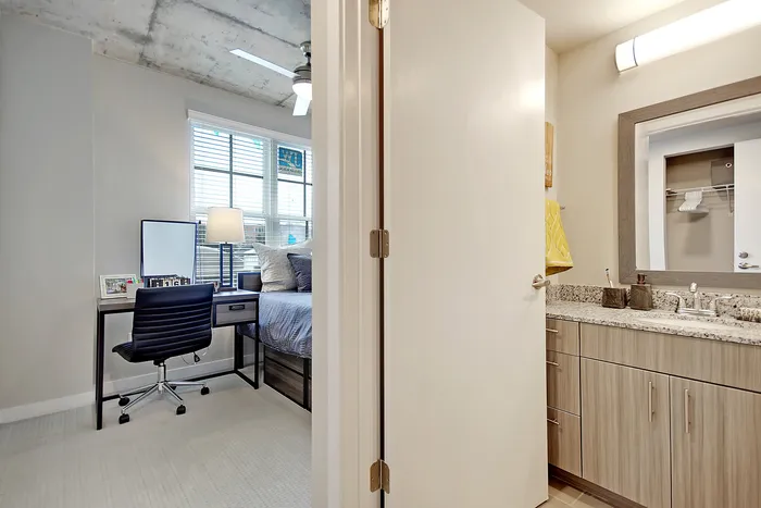 Modern apartment with a cozy bedroom and sleek bathroom, featuring a stylish desk by a window and a granite-topped vanity.