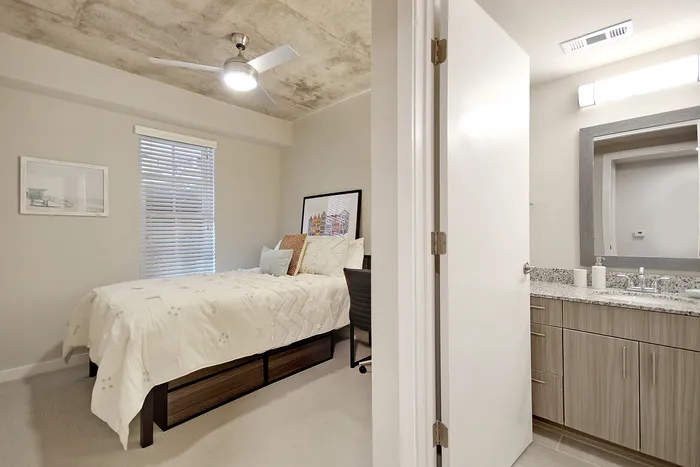 Modern bedroom with stylish ceiling fan, large window, and adjacent bathroom featuring elegant granite countertops.