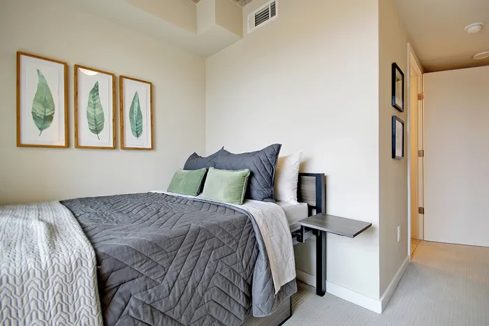 Cozy bedroom with a modern aesthetic, featuring a plush bed with gray and green pillows, framed leaf art, and neutral walls.