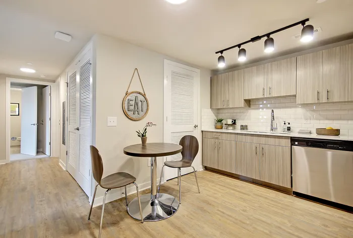 Modern kitchen with sleek cabinetry, stainless steel dishwasher, and cozy dining area featuring a circular table and two chairs, enhanced by bright track lighting.