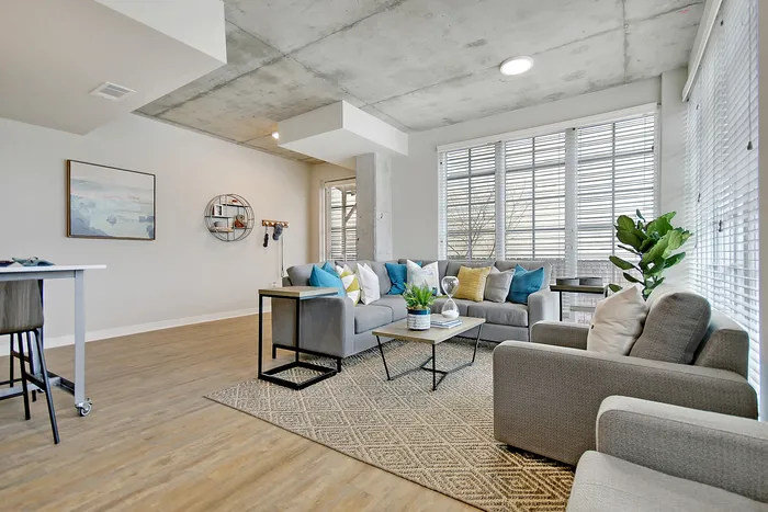 Modern living room with stylish gray furniture, vibrant cushions, and large windows offering abundant natural light.