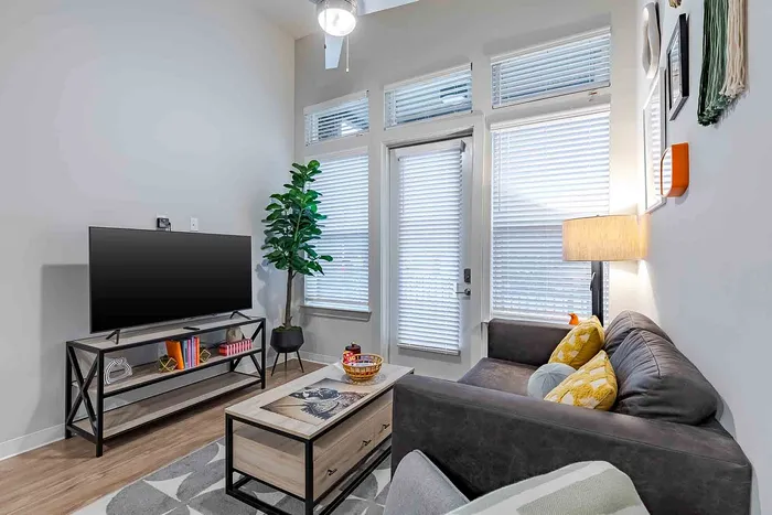 Bright and stylish living room with modern decor, featuring a cozy grey couch, vibrant cushions, and a sleek TV setup beneath large windows.