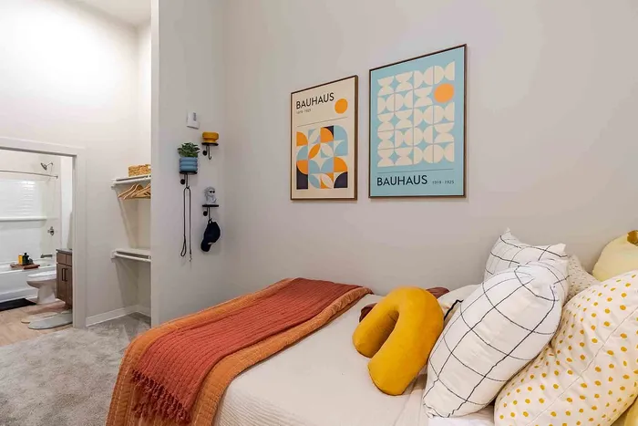 Cozy bedroom with modern decor featuring Bauhaus posters, a plush bed with vibrant pillows, and a glimpse into a stylish bathroom.