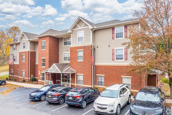 Charming brick apartment building with modern accents, surrounded by autumn foliage, ample parking space, and a welcoming entrance.