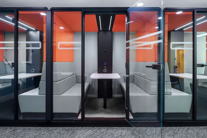 Modern and vibrant office pod with sleek glass walls, cozy seating, and bright orange accents ideal for private meetings.