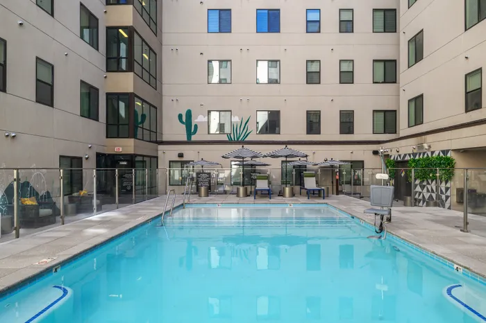 Modern apartment courtyard featuring a sparkling swimming pool surrounded by stylish seating areas and elegant umbrellas, with contemporary wall art and large windows.