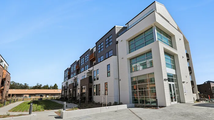Modern multi-story apartment complex with sleek architectural design and large windows, surrounded by well-maintained landscaping under a clear blue sky.