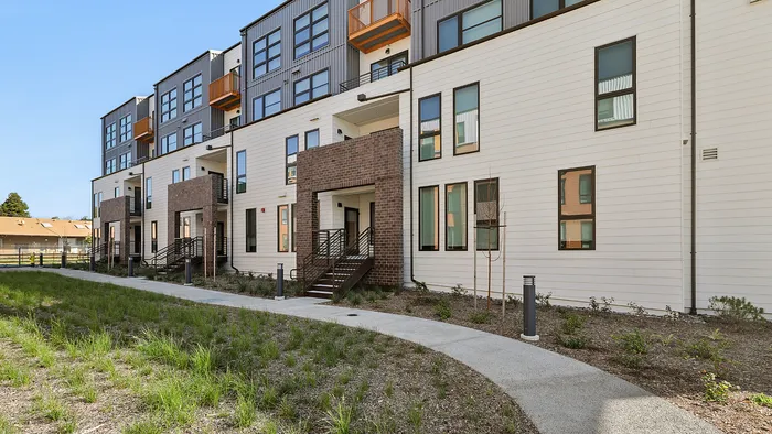 Modern apartment building exterior with sleek design, multiple floors, and large windows, surrounded by landscaped grounds and a well-maintained path.
