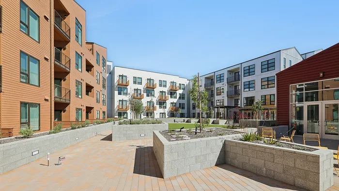 Chic modern apartment complex featuring multi-story buildings with balconies, surrounded by a spacious courtyard garden and stylish seating under a clear sky.