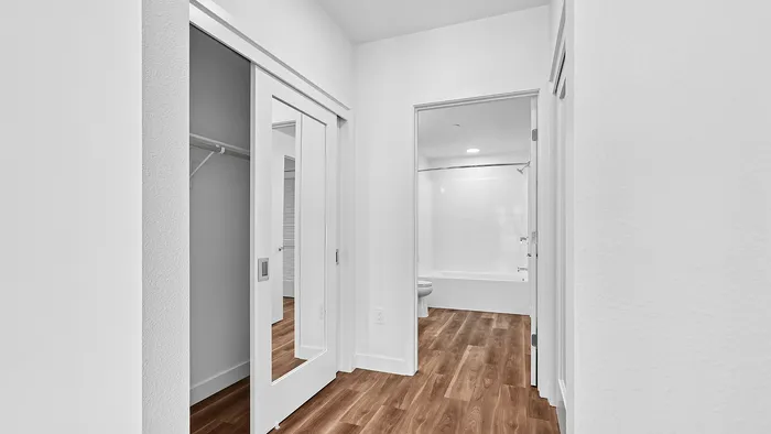 Contemporary hallway with sleek wooden flooring, mirrored closet doors, and a bright modern bathroom.