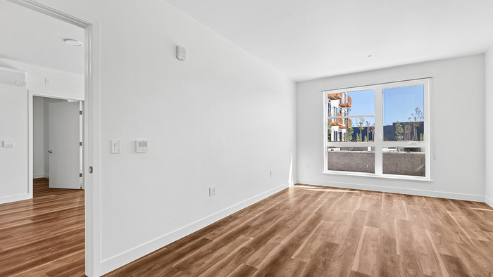 Bright, empty room with elegant hardwood flooring, large window offering abundant natural light, and a view of nearby architecture.