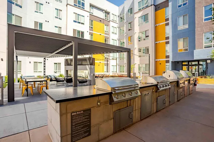 Modern apartment courtyard featuring outdoor BBQ grills and stylish seating under a chic pergola, surrounded by colorful building facade.