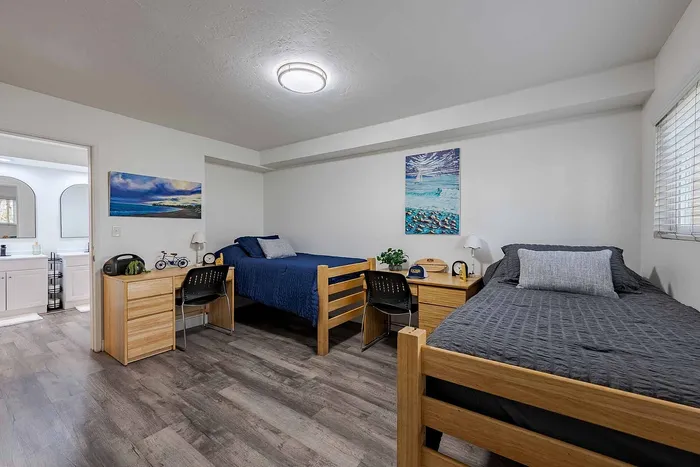 Modern shared bedroom with stylish wood furniture, featuring twin beds, sleek desks, and vibrant coastal artwork, complemented by an en-suite bathroom.