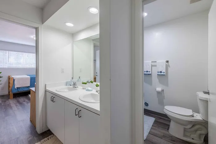 Modern apartment bathroom showcasing a dual-sink vanity and pristine fixtures, leading to a cozy bedroom with ample natural light.